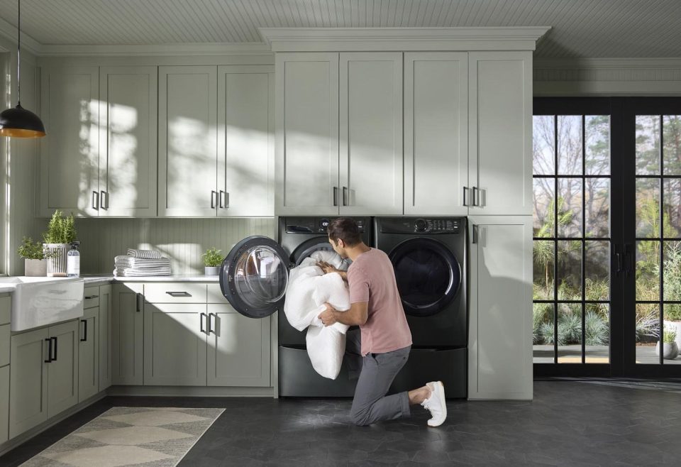 A person kneels in front of a black washing machine, loading white towels. The laundry room has light green cabinets, potted plants, and large windows showing an outdoor view. The floor is dark-colored and the walls are pale.