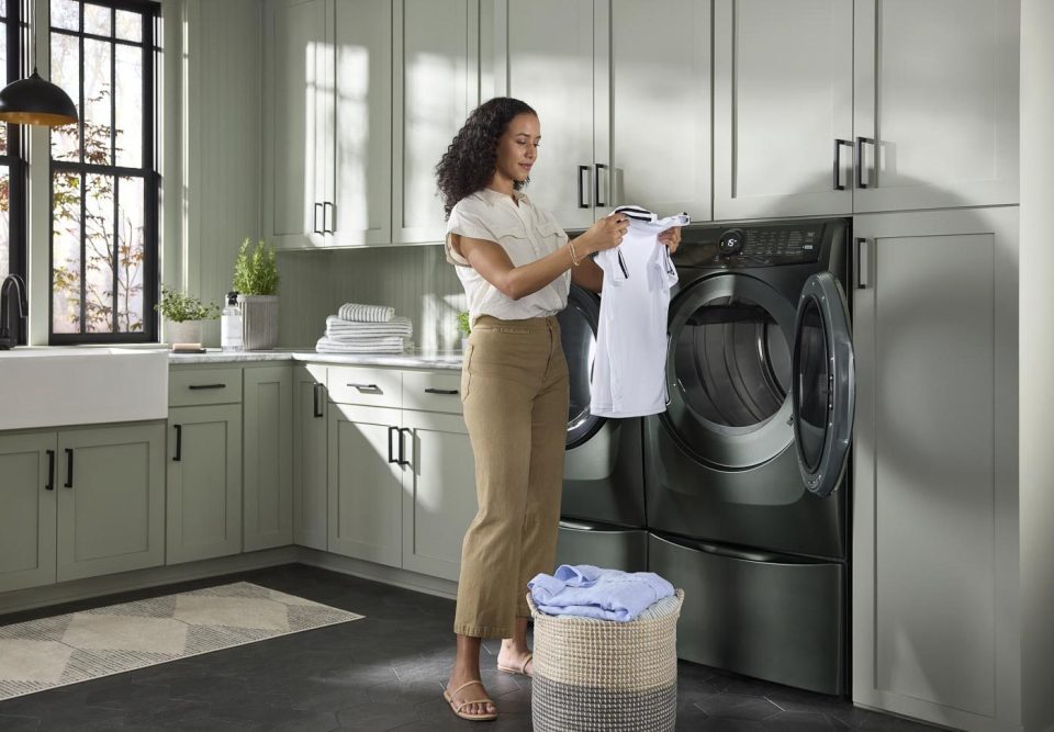 A person is folding a white shirt in a laundry room with green cabinets. A front-loading washer and dryer are next to them, and a basket with clothes is on the floor. Sunlight comes through a large window, illuminating the tidy space.