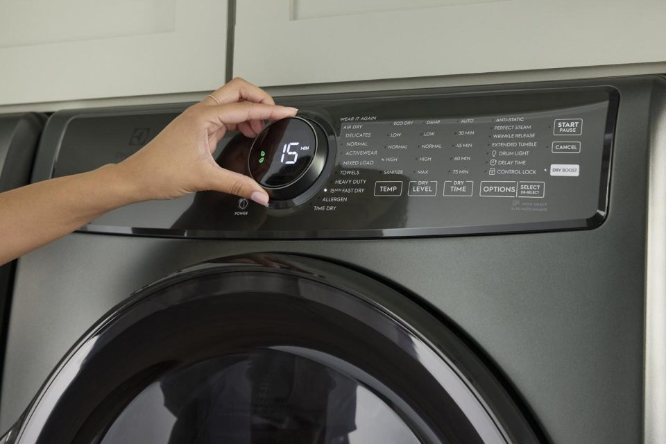 A person adjusts the settings on a modern washing machine. The display shows "15" and various options are visible, including wash cycle types and start button. The machine is silver with a digital control panel.