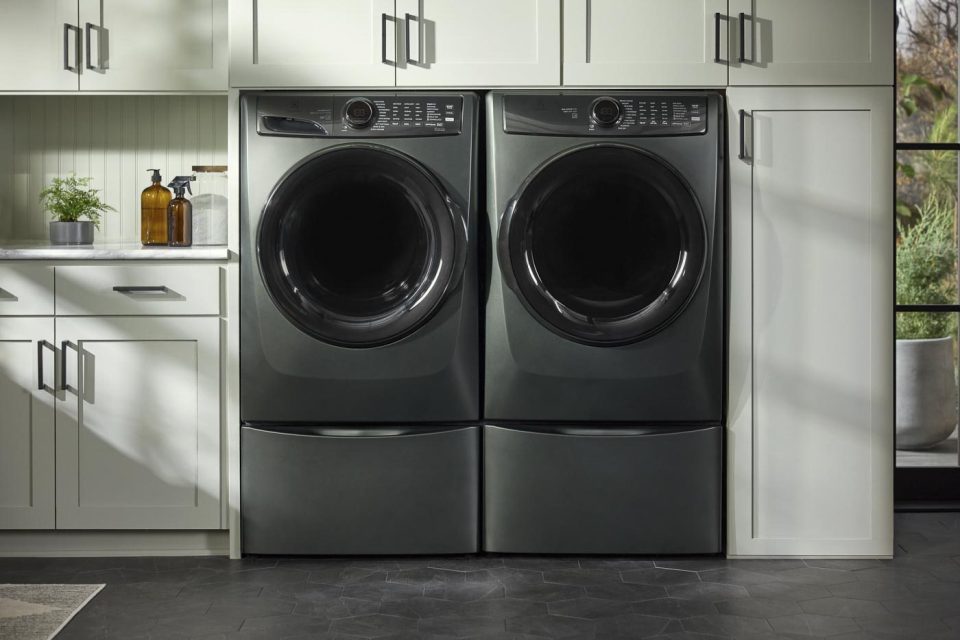 A laundry room features a modern washer and dryer set in a sleek, matte black finish. They are built into cabinetry with grey cabinets and a countertop. A plant and soap dispenser are on the counter, and a window brings in natural light.