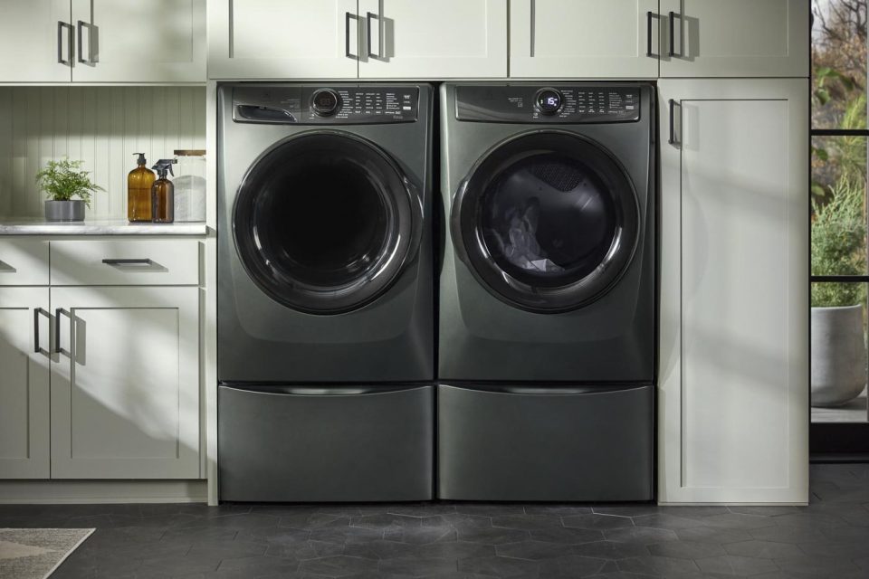 A modern laundry room features a black front-loading washer and dryer set, neatly installed between pale green cabinets. A countertop with plants and soap dispenser is visible on the left, and the room has a dark tile floor.