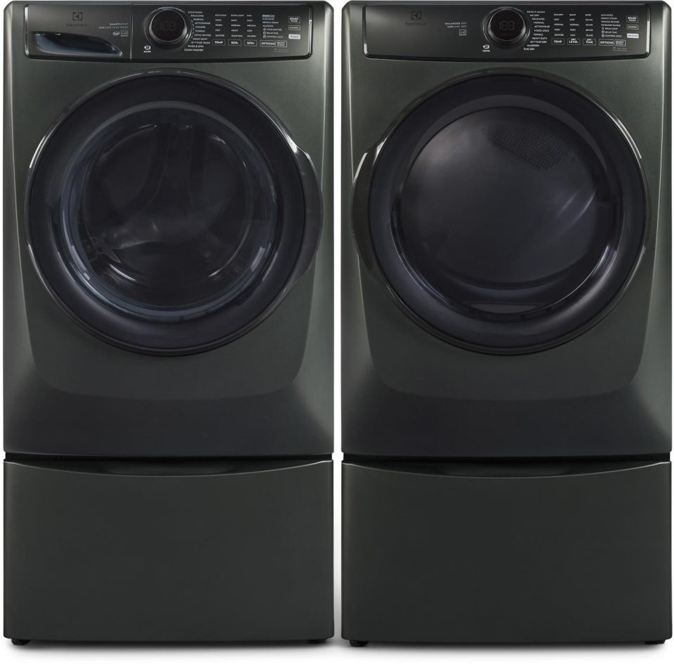 A pair of modern dark gray front-loading laundry machines, a washer on the left and a dryer on the right. Both feature digital displays and sit atop storage pedestals.