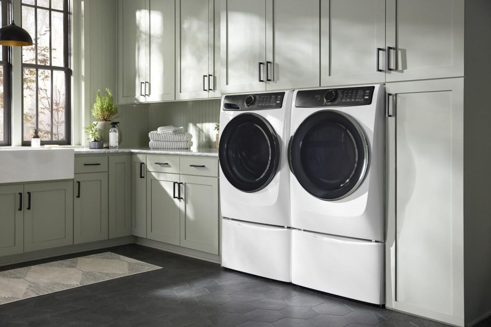 A modern laundry room with light green cabinets and black hardware. A front-loading washer and dryer set is placed side by side. A stack of folded towels and a plant are on the countertop, and a striped rug sits on the dark herringbone floor.