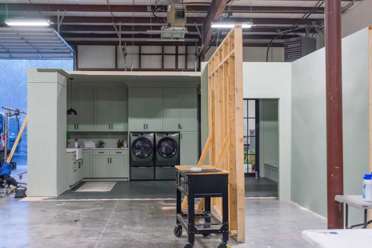 A partially constructed set in a warehouse features light green walls, a kitchen area, and a laundry section with a washer and dryer. An unfinished wooden frame stands in the foreground, and a cart is nearby. Overhead lighting and a garage door are visible.