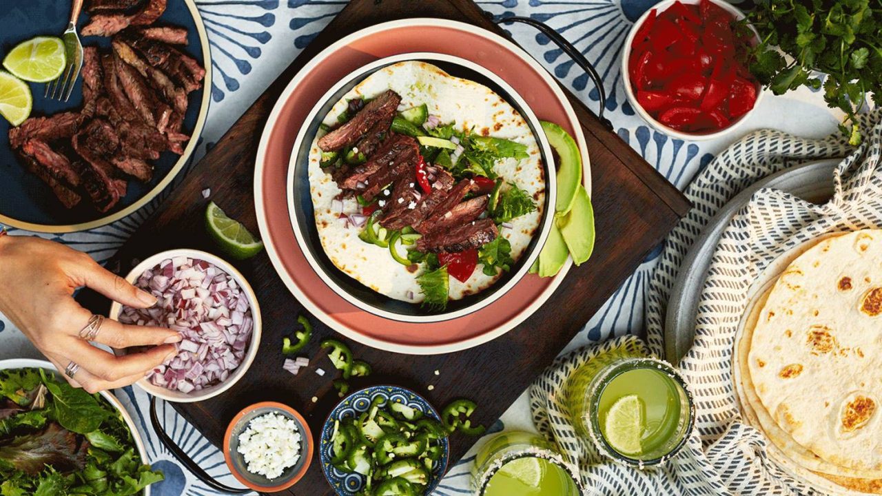 Flat lay photograph of skirt steak tacos and flour tortillas with bowls of colorful toppings.