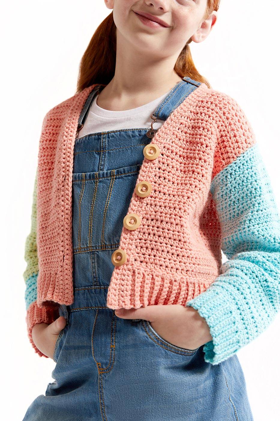 A child wearing a pink and blue crocheted cardigan over a denim dress with overall straps. The cardigan has wooden buttons and colorful sleeves. The child stands confidently against a plain white background.