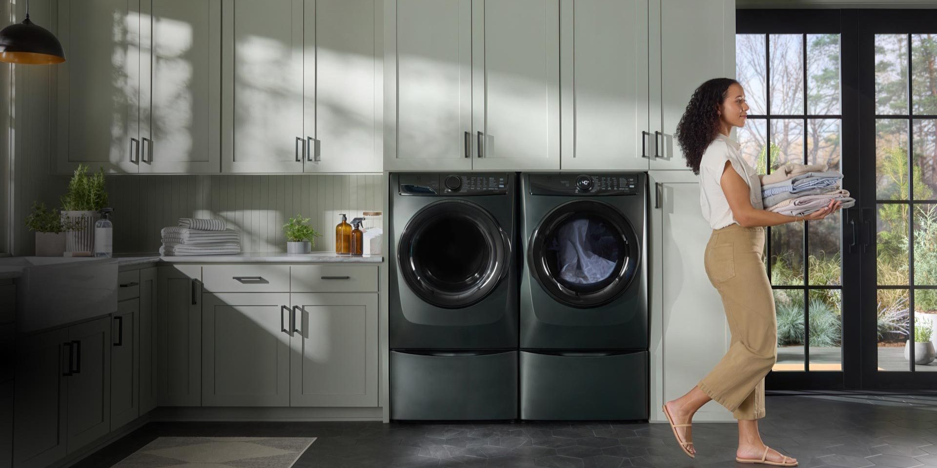 A woman carries folded laundry in a modern laundry room with dark green cabinets, a large window, and a front-loading washer and dryer. Sunlight streams in, illuminating plants and a detergent bottle on the countertop.