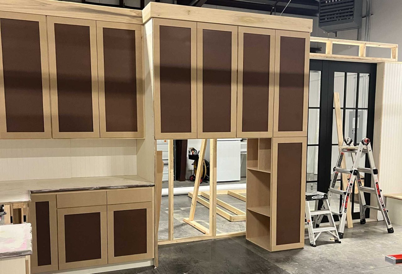 A kitchen under construction with light wood cabinets and dark brown panels. Two ladders are positioned near the cabinets, and construction materials are scattered around. A set of black-framed glass doors is visible in the background.