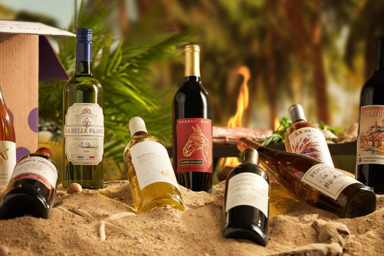 Image of wine bottles propped in the sand on the beach in the evening with palm trees in the background.