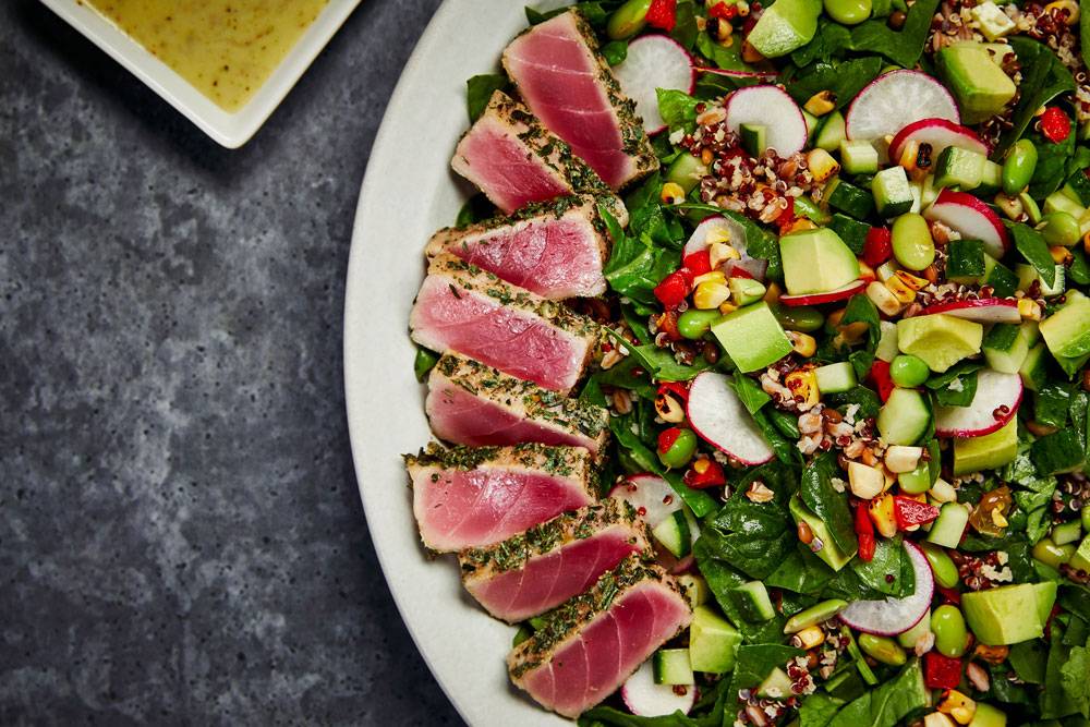 A plate of seared tuna slices is arranged next to a colorful salad with leafy greens, quinoa, radishes, edamame, avocado, and corn. A small bowl of dressing is visible in the corner on a textured gray surface.