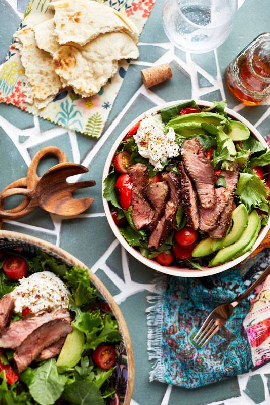 Flat lay photograph of a plate of grilled loin strip streak and a bowl of salad set on a bright blue tablecloth.