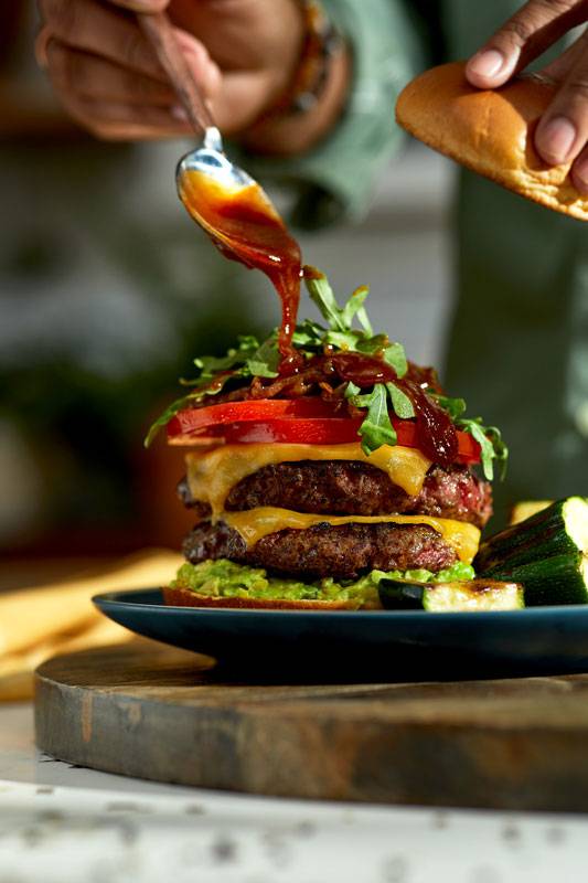 Close-up photograph of double cheeseburger with the chef's hands drizzling sauce on top before placing the top bun.