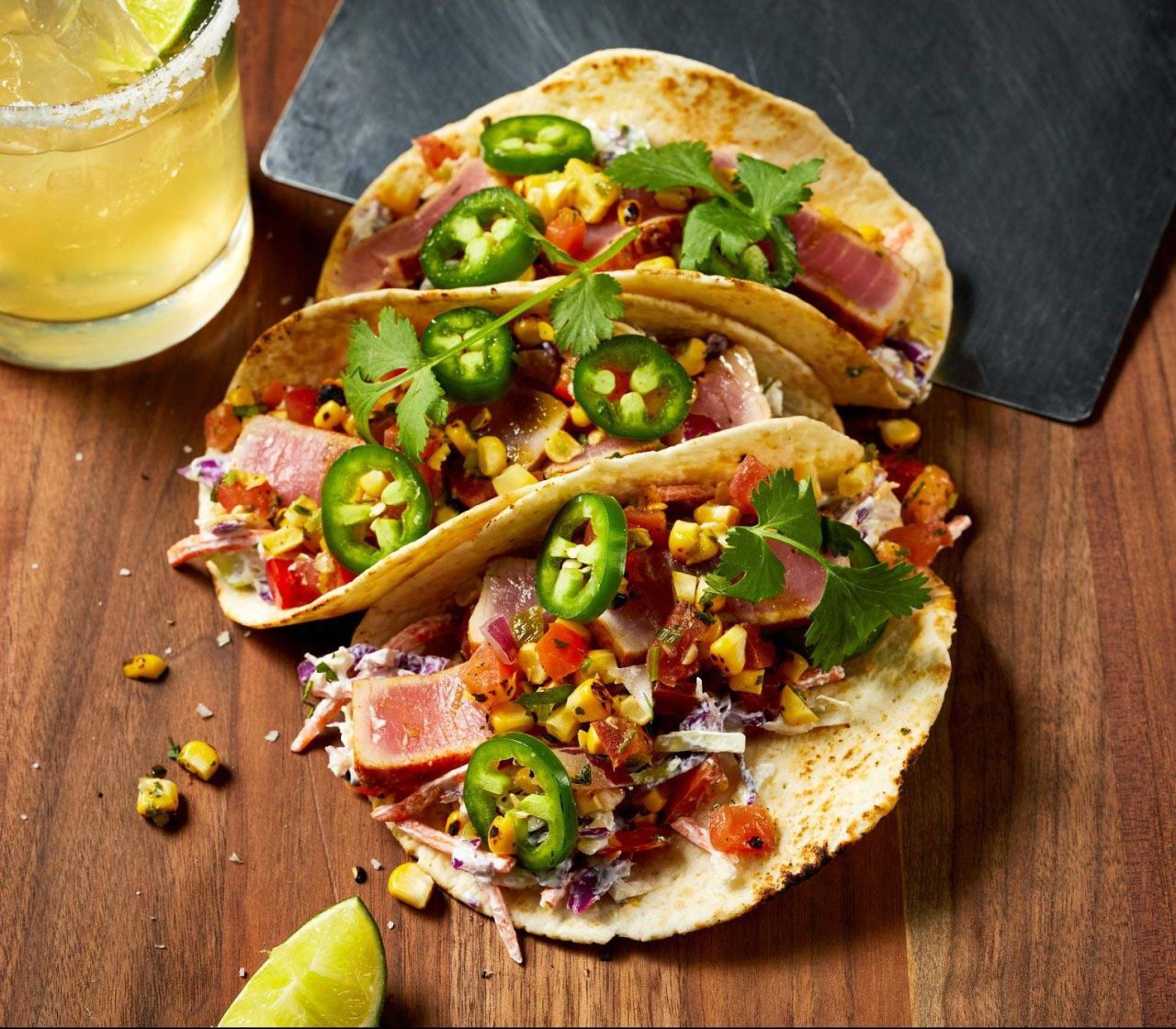 Three tacos filled with seared tuna, sliced jalapeños, cilantro, diced tomatoes, and charred corn, served on a wooden table with a glass of margarita and a lime wedge on the side.