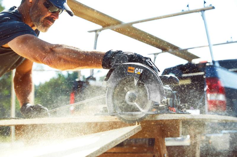 A person wearing a cap, sunglasses, and gloves operates a circular saw, cutting through a wooden board outdoors. Sawdust flies around, and a truck is parked in the background on a sunny day.