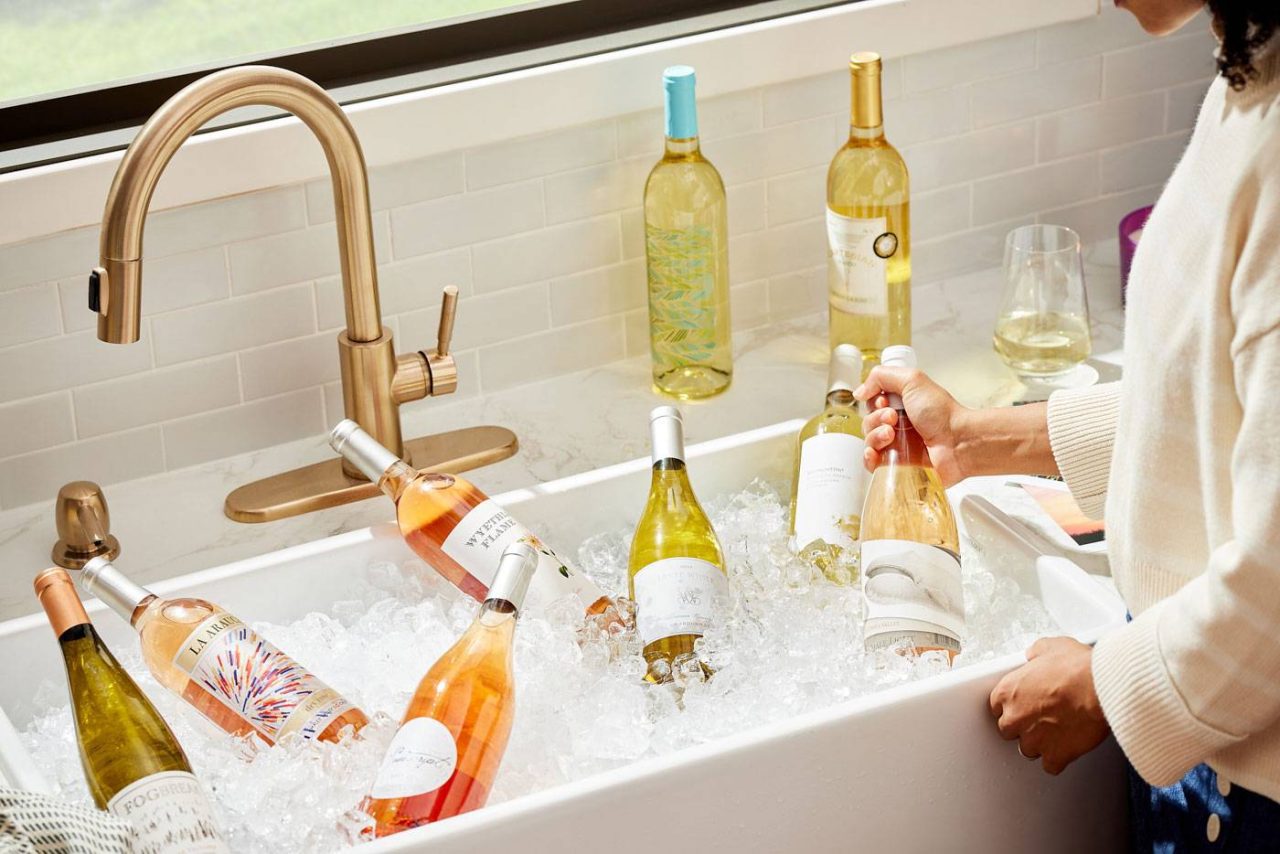 A person is placing a wine bottle in a sink filled with ice, surrounded by several other chilled wine bottles. A glass of white wine and additional bottles are on the countertop near a window.