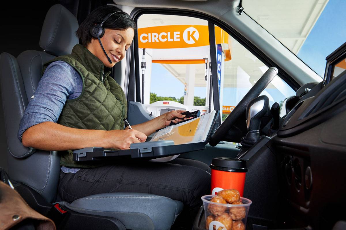 Commercial photography of a person wearing a headset is sitting in the driver's seat of a vehicle, using a clipboard to write. A Circle K gas station is visible outside. There is a cup of coffee and a container of snacks on the console.