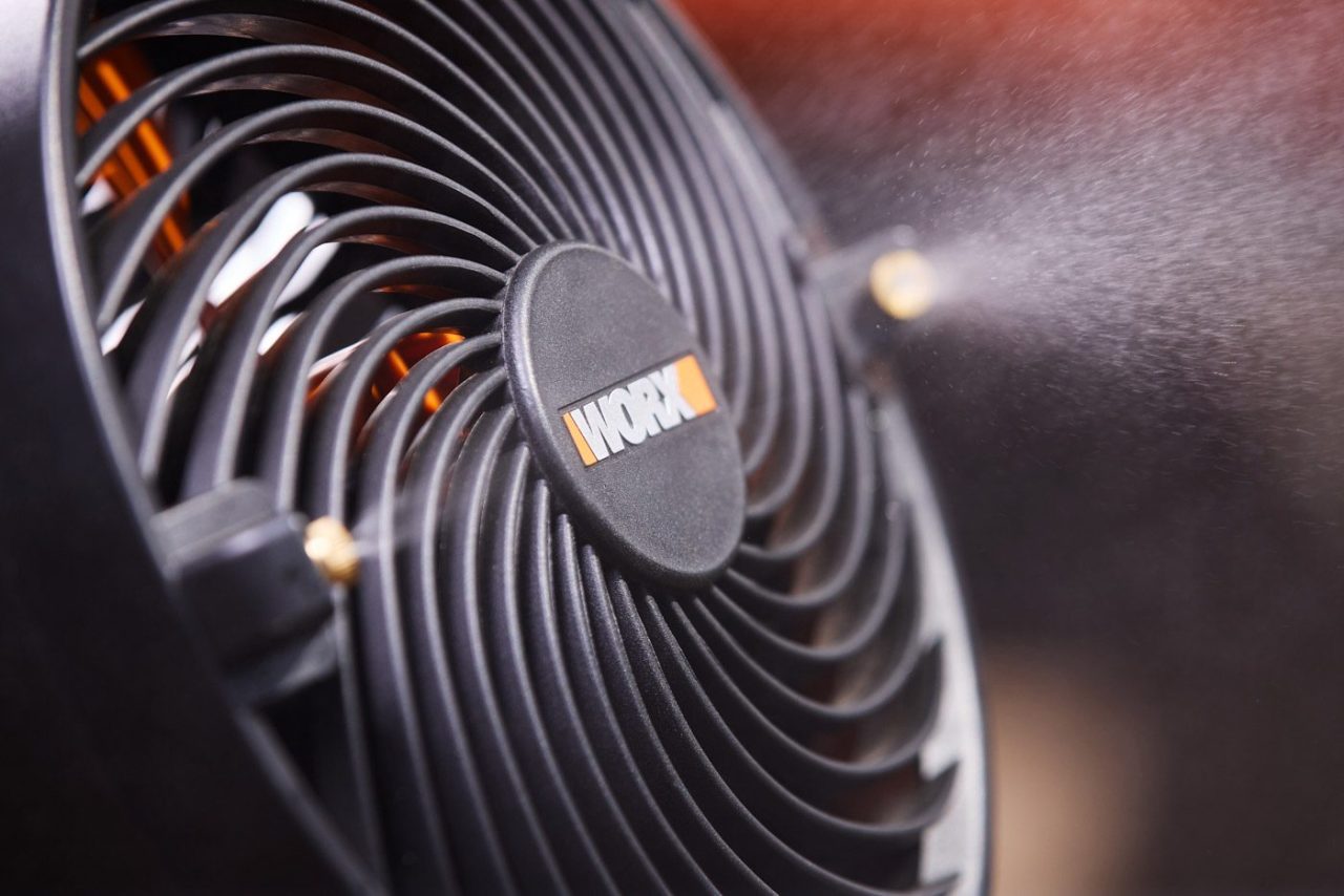 Close-up of a black fan with a protective grille, branded with the logo "Worx" at the center. The fan is in motion, creating a visible mist or steam dispersing through its blades, against a blurred background.