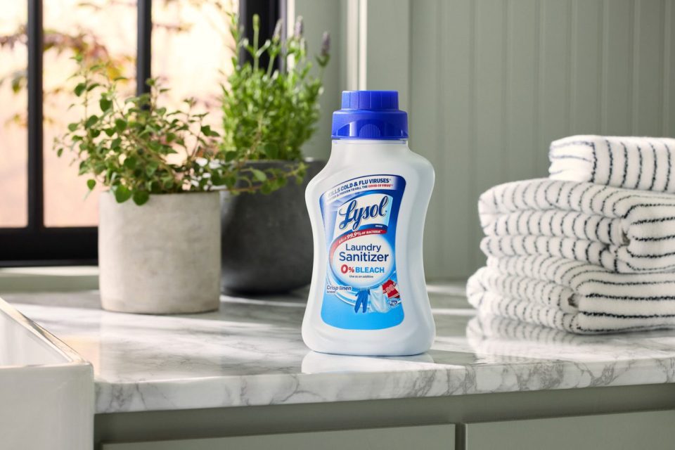 A white and blue bottle of Lysol Laundry Sanitizer sits on a marble countertop. Nearby, there are folded striped towels, potted plants, and a window with black frames, creating a bright and clean laundry room atmosphere.