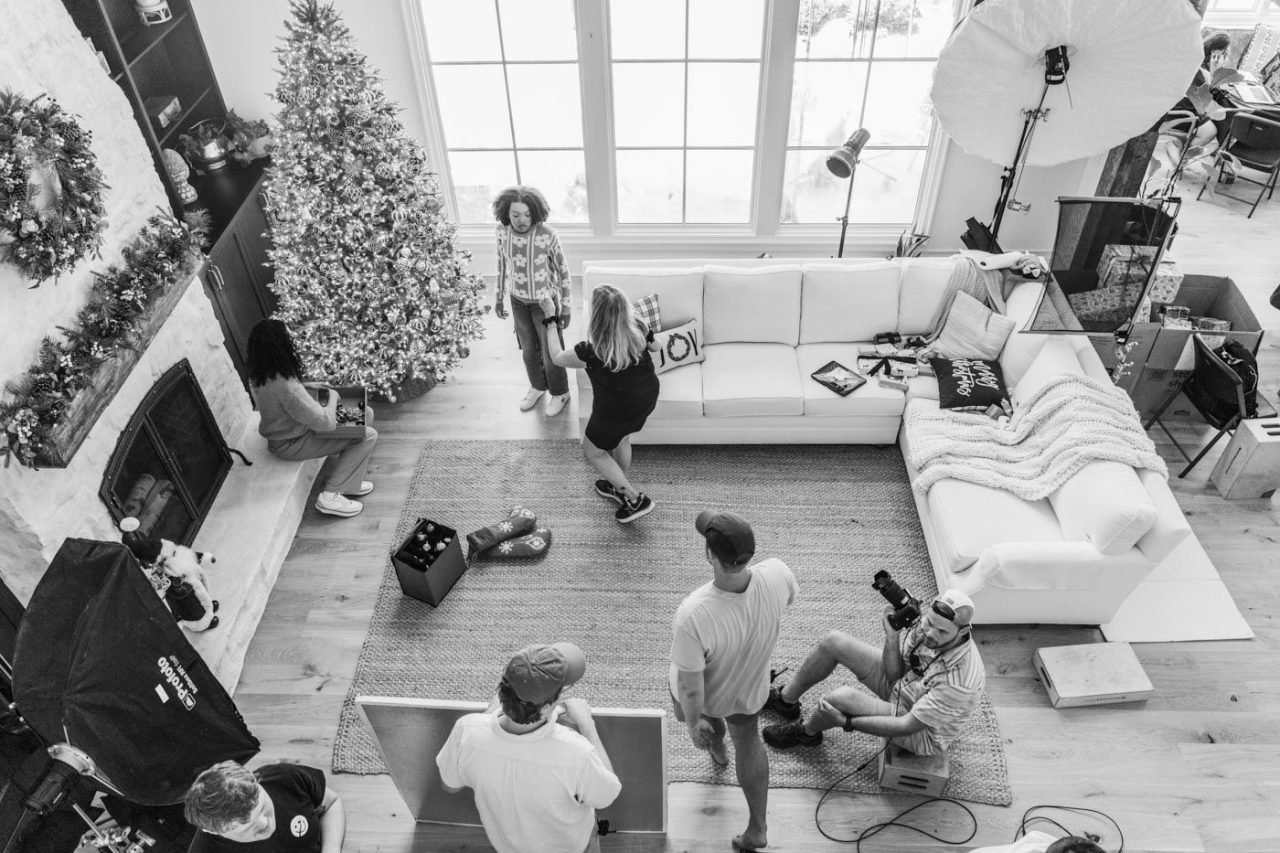 A black-and-white image reveals a living room transformed into a holiday-themed production studio. People are arranging a photo shoot near the Christmas tree, with equipment and lights strategically set up around a sofa on the carpeted floor, as several individuals fine-tune props and cameras.