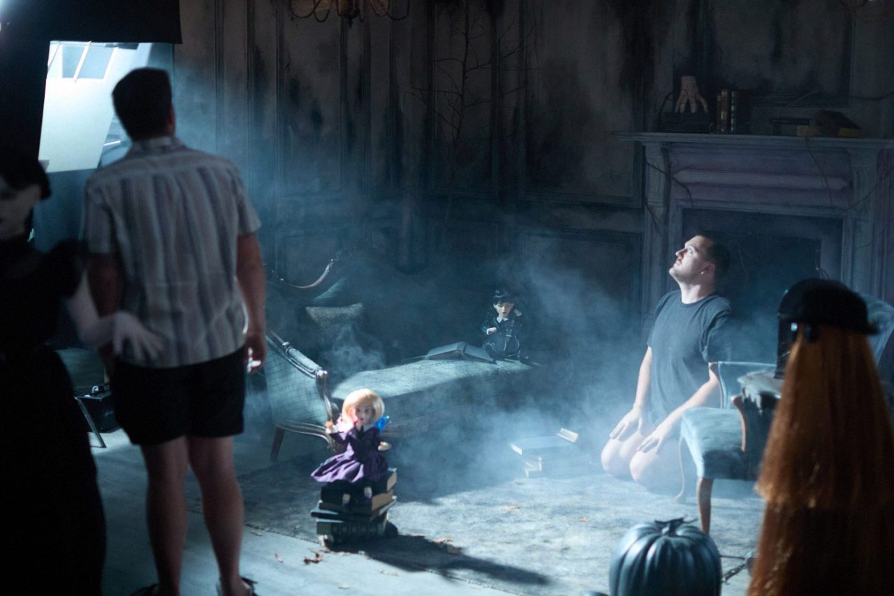 A foggy room with a man kneeling, facing a dimly lit area. Dolls and Halloween props surround the scene. An armchair, a fireplace, and eerie decorations fill the background, creating a haunting atmosphere. People are partially visible on the set.