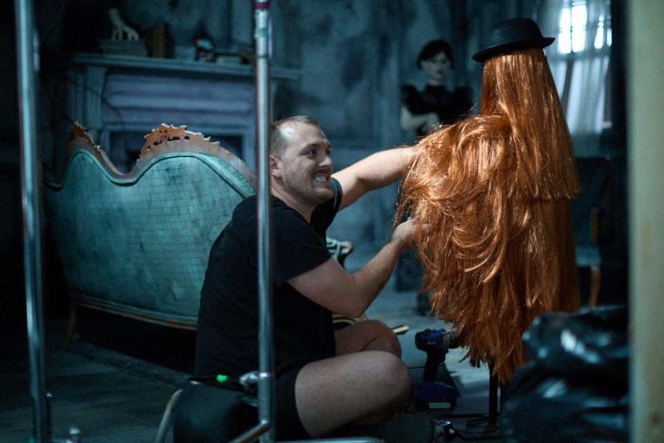 A man wearing a black shirt kneels on the floor, styling a long wig on a mannequin head. The room has dim lighting, a vintage couch, and a figure in the background. The atmosphere appears to be a film or theater production set.