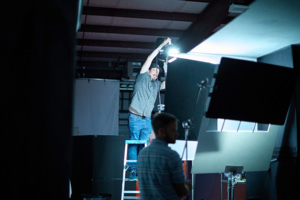 A man stands on a ladder adjusting a light in a dimly lit studio. In the foreground, another person in a striped shirt appears blurred next to professional lighting equipment and screens. The setting suggests a photo or video shoot.