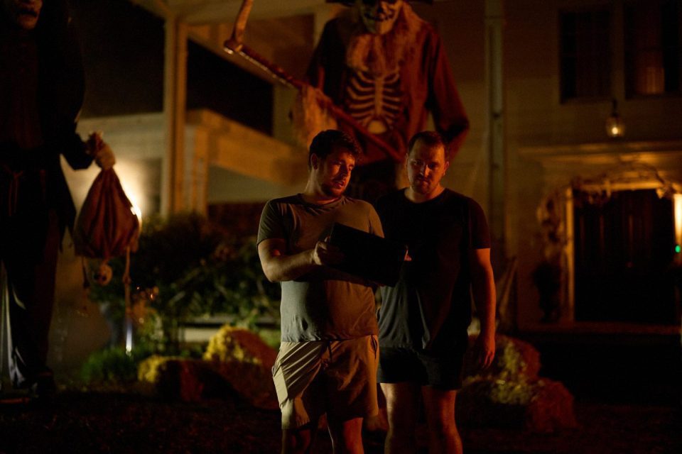 Two people stand outside at night, looking at a tablet. Behind them, a large Halloween skeleton decoration with a scythe looms. The scene is dimly lit, creating a spooky atmosphere.