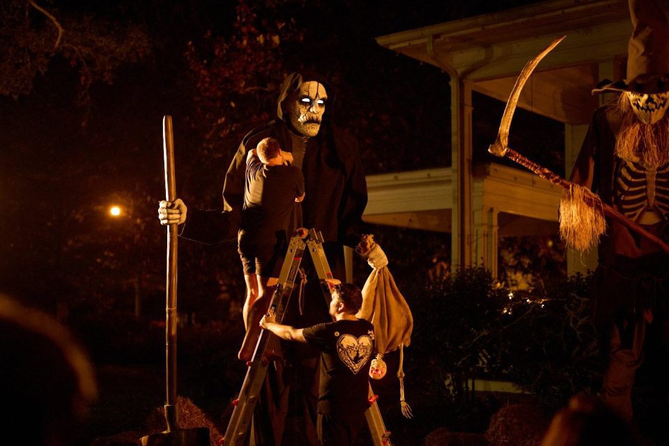 A person on a ladder decorates a tall, spooky Grim Reaper figure holding a shovel, illuminated by warm orange lights in a nighttime setting. Another figure, resembling a pumpkin scarecrow with a scythe, stands nearby.