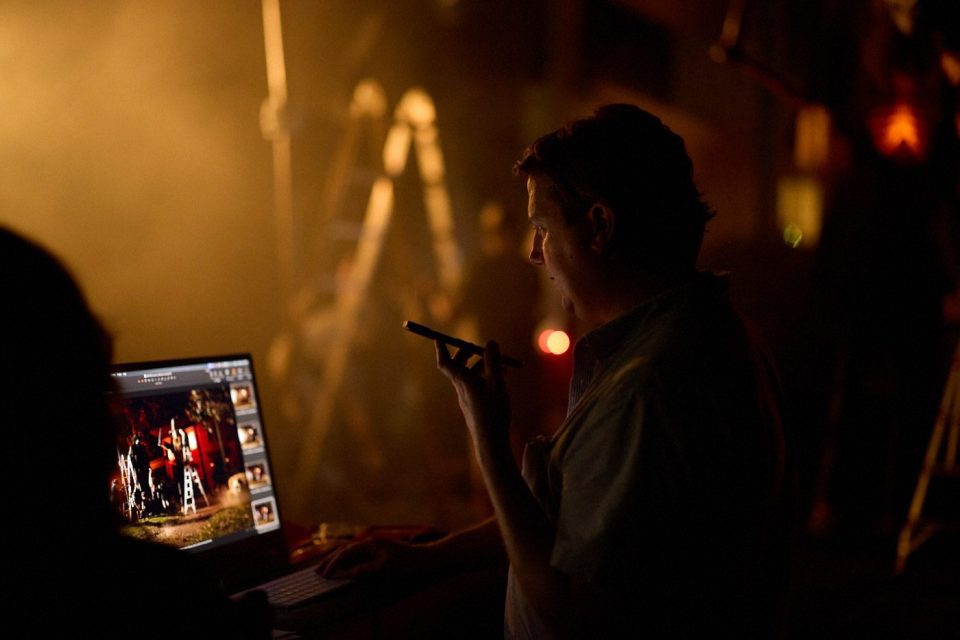 A person holds a pen and looks at a laptop screen displaying images. The scene is dimly lit with warm lighting, creating a dramatic atmosphere. A blurred figure and a ladder are visible in the background.