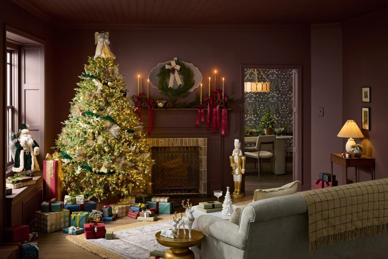 A cozy living room decorated for Christmas. A large, lit Christmas tree adorned with ornaments stands beside a fireplace with candles and a wreath. Gifts are scattered around the tree, and festive decorations fill the room, creating a warm atmosphere.