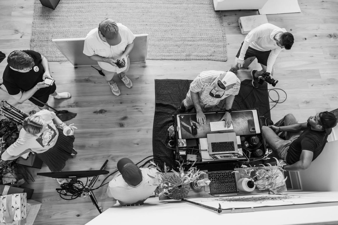 A black and white overhead shot of a creative team working together. Six people are gathered around a table with laptops and cameras, discussing and collaborating. The setting appears casual, with a wooden floor and scattered objects.