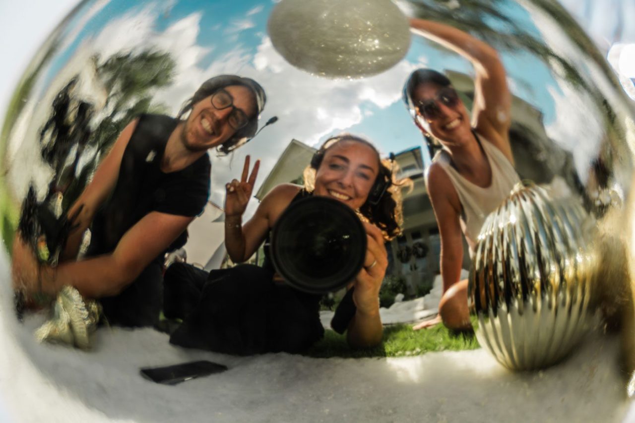 Three people smiling at their reflection in a metallic ball. One person holds a camera and flashes a peace sign. The scene is outdoors with trees and a building in the background, under a clear blue sky.