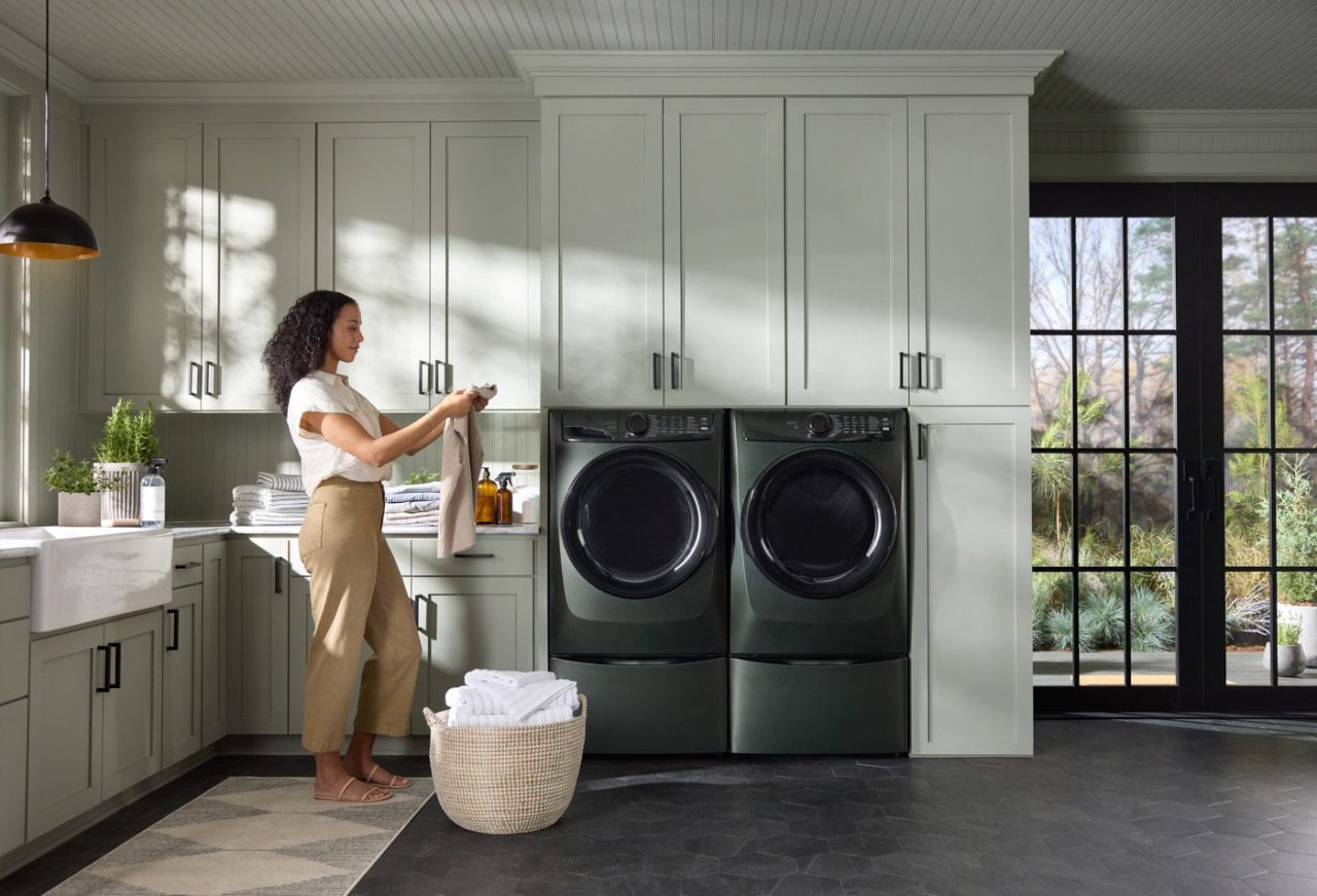 A woman stands in a modern laundry room with green cabinets, placing clothes into a washing machine, capturing the essence of lifestyle photography. A basket of laundry is on the floor, while large windows offer natural light and a picturesque view of the garden.