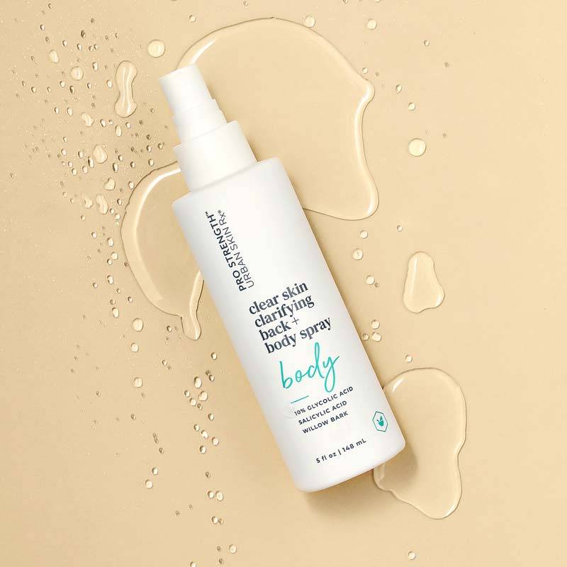 A spray bottle of "Pro Strength Clearskin Clarifying Back & Body Spray" rests on a beige surface adorned with water droplets, captured in stunning product photography. The label emphasizes 10% glycolic acid and willow bark, with the bottle's white design featuring teal and black text.