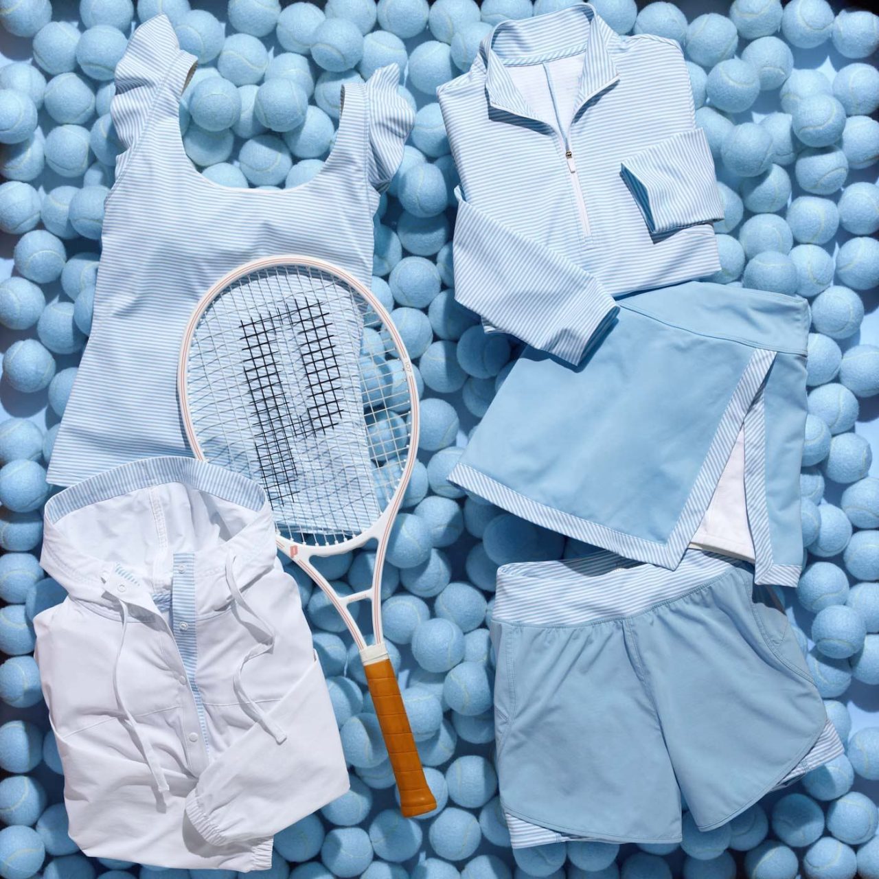 A collection of blue and white tennis apparel, including a sleeveless top, zip-up jacket, shorts, skirt, and collared shirt, arranged on a pile of light blue tennis balls. A white tennis racket with a wooden handle lies across the clothes.