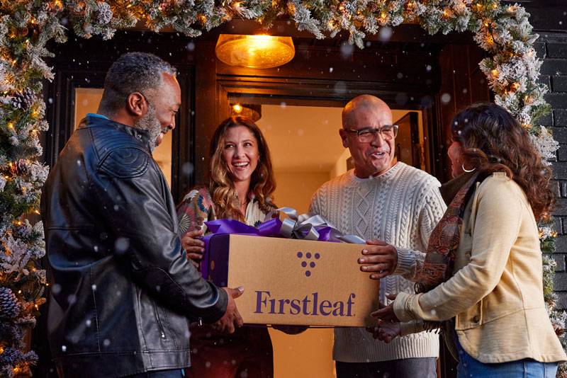 Four people stand in a snowy doorway decorated with festive garlands. Two are handing a Firstleaf box with a purple ribbon to the other two, all smiling and engaged in a cheerful holiday exchange.