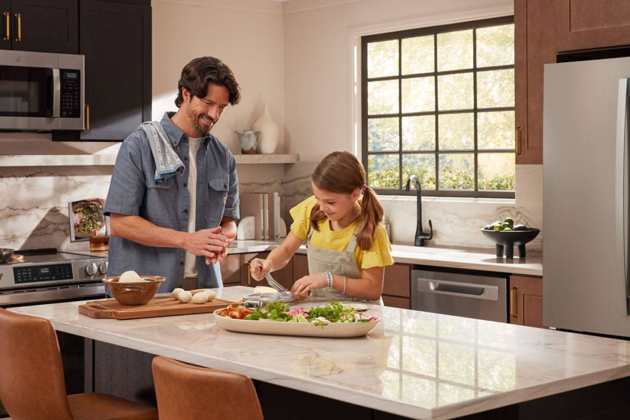 A man and a young girl are cooking together in a modern kitchen. The man is smiling while using a pepper grinder, and the girl is chopping vegetables. The countertop is decorated with various kitchen items and fresh ingredients.