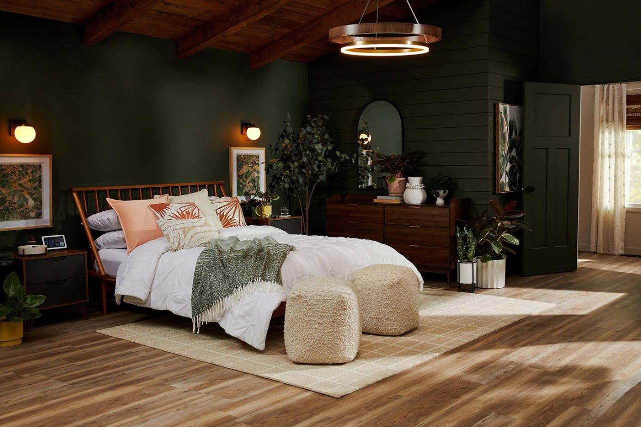 A cozy bedroom with a wooden bed, white bedding, and green accent wall. Two plush ottomans are in front of the bed on a light rug. Wooden furniture and decorative plants enhance the space, lit by modern lighting.