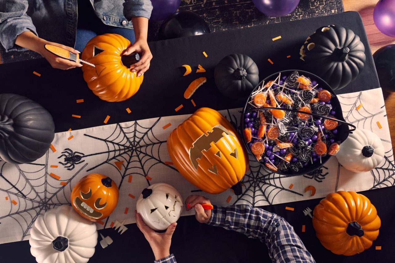 People carving pumpkins at a table decorated with Halloween-themed items, including a spiderweb runner and a bowl of treats. Several carved pumpkins and cutting tools are visible, as well as black, white, and orange pumpkins.