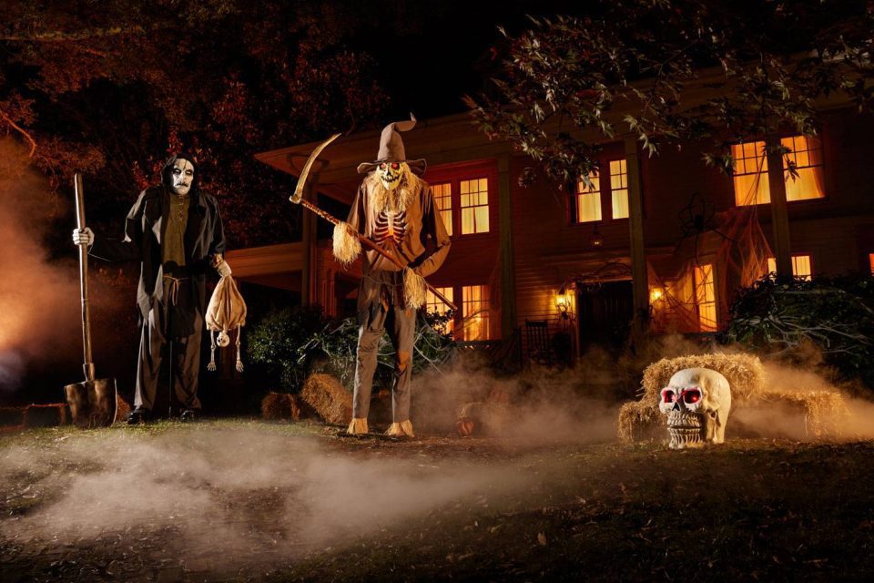 A spooky Halloween scene featuring two creepy figures with masks and costumes holding a spade and scythe. A large skull with red eyes is on the foggy ground, set against a dimly lit house decorated for the holiday.