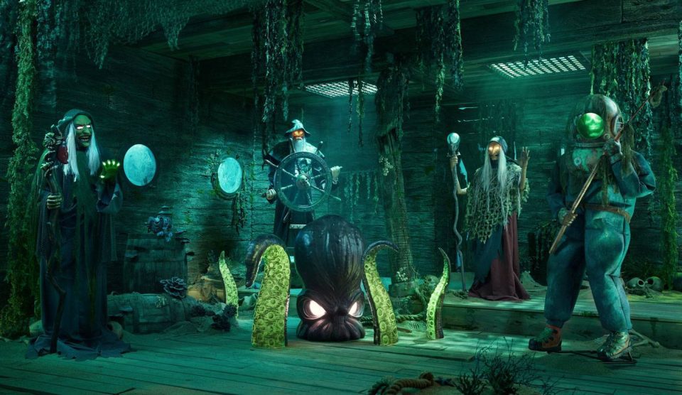 Four people in eerie costumes stand in a dimly lit, overgrown room with a nautical theme. An octopus emerges from the floor. They hold various props: a globe, a ship wheel, and a candelabra. Greenish lighting adds to the eerie ambiance.