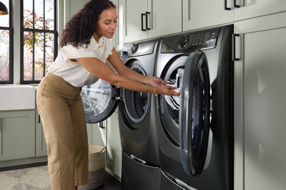 A woman, depicted in lifestyle photography, is loading clothes into a modern front-loading washing machine in a laundry room with green cabinets. Clad in a white blouse and beige pants, she stands beside a dryer as sunlight streams through the window.