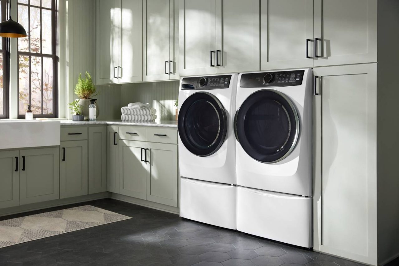 A modern laundry room with light green cabinets, a farmhouse sink, and a window streaming in natural light. The lifestyle photography perfectly captures the stacked front-loading washer and dryer units, neatly folded towels on the counter, and a rug gracing the dark tiled floor.