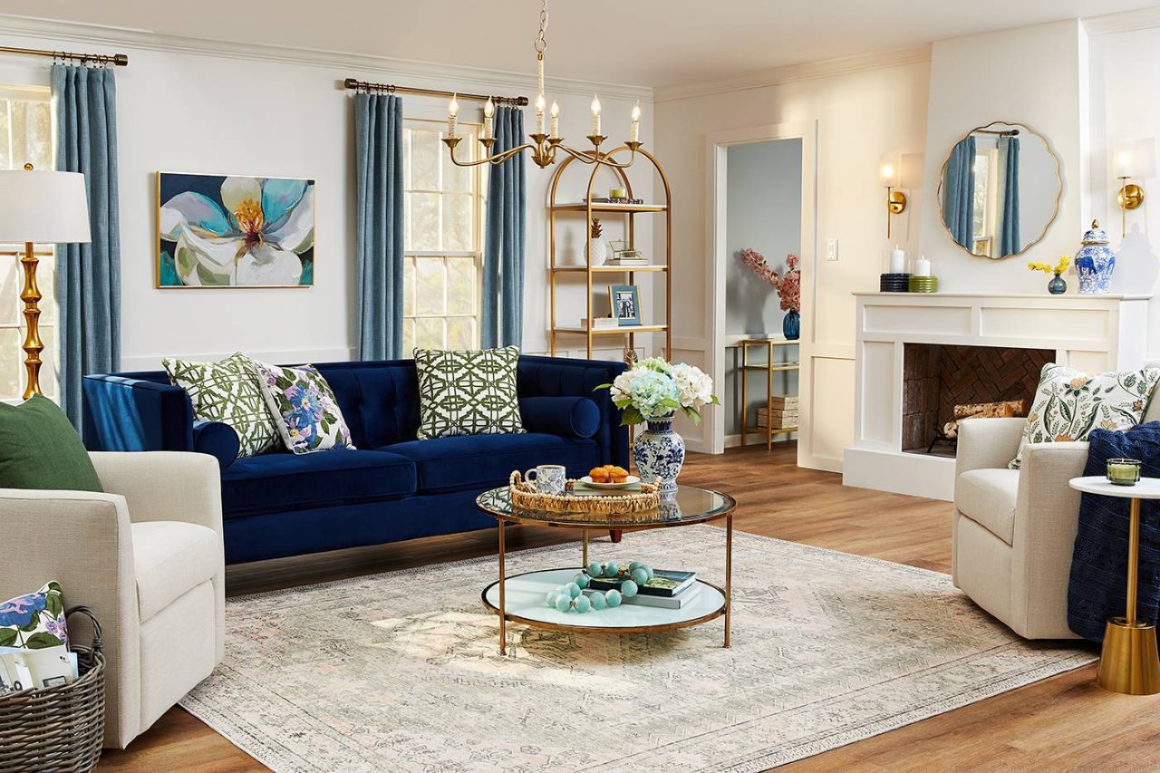 A cozy living room perfect for lifestyle photography, featuring a blue velvet sofa, patterned pillows, and a glass coffee table with flowers. Beige armchairs and a rug adorn the hardwood floor, while a fireplace, round mirror, and decorative shelves complete the elegant setting.