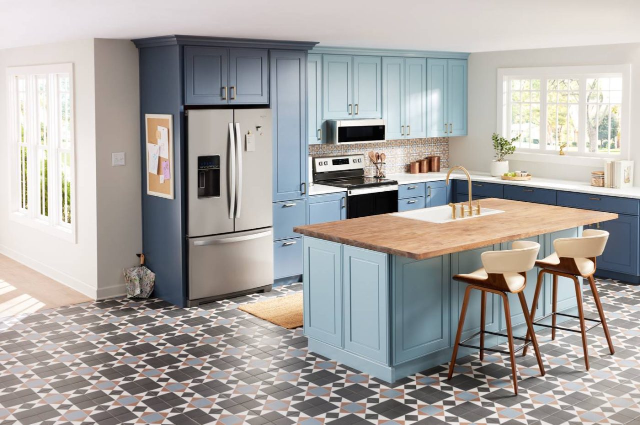 A modern kitchen with blue cabinets, stainless steel appliances, and a wooden island with two tall chairs. The floor's geometric black, white, and pink tile pattern sets the scene perfectly for lifestyle photography. Large windows flood the space with natural light.