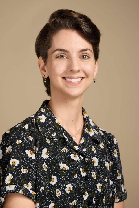 Meet the team: A person with short brown hair smiles at the camera, wearing a black shirt adorned with a white daisy pattern and small earrings. The background is a neutral beige color, perfectly highlighting their friendly demeanor.