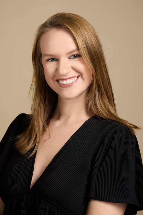 Meet the team: A smiling woman with long, light brown hair wears a black V-neck top. She poses against a neutral beige background, looking directly at the camera.