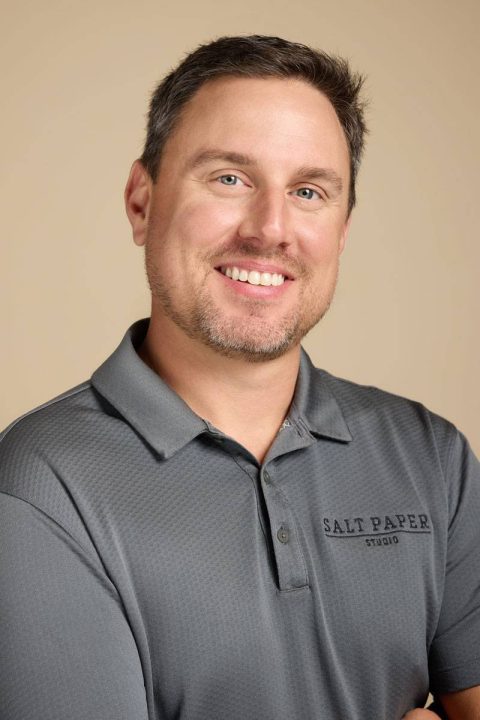 Meet the team: A man with short brown hair and a beard, wearing a gray polo shirt embroidered with "Salt Paper Studio," smiles confidently, arms crossed against a beige background.