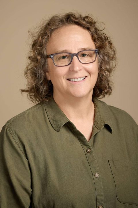 Meet the team: A person with curly light brown hair, wearing glasses and a green button-up shirt, smiles warmly against a beige background.