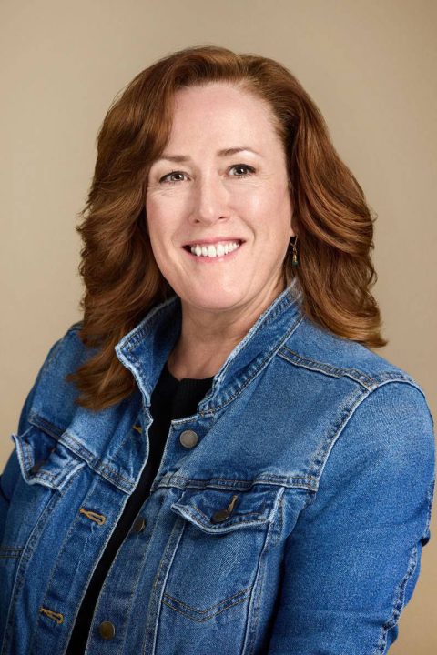 Meet the team: A woman with shoulder-length auburn hair smiles warmly at the camera, wearing a blue denim jacket over a black top. The backdrop is a neutral beige, highlighting her bright demeanor.